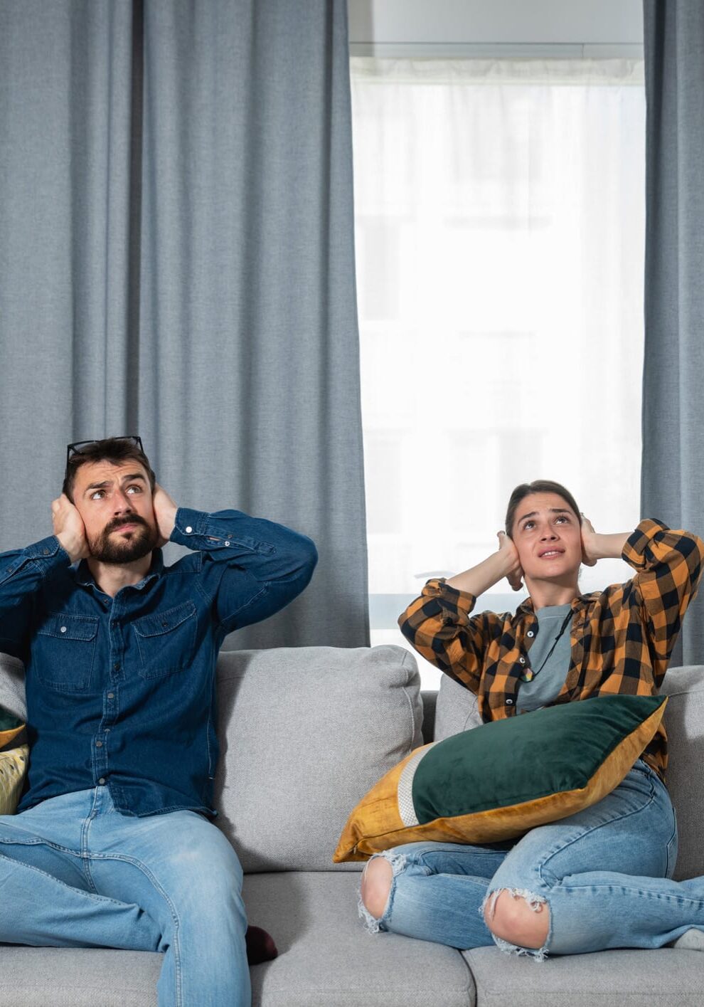 Young couple is sitting on a sofa in their apartment looking up and holding their hands to plug their ears as a neighbour upstairs is having a party and playing loud music or renovating the apartment